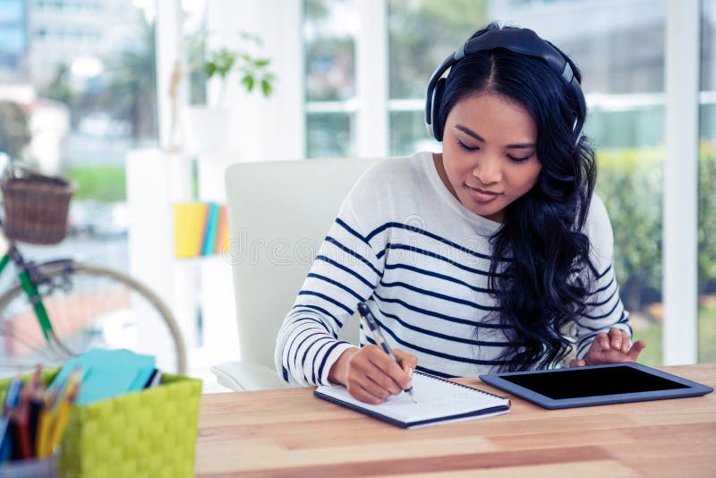Mujer asiática sonriente con los auriculares que escribe en la libreta imagen de archivo libre de regalías Mujer asiática sonriente con los auriculares que escribe en la libreta imagen de archivo libre de regalías