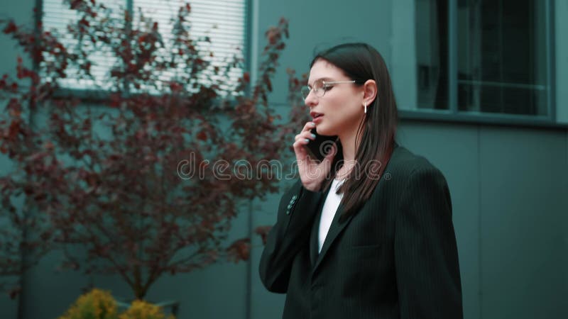 Mujer asiática sonriente con traje formal usando teléfonos móviles escribiendo mensajes de texto caminando con una laptop fuera de