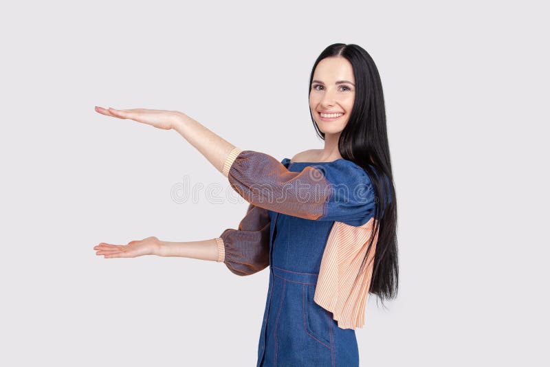 Mujer atractiva alegre joven en el vestido azul claro de los vaqueros que muestra algo grande con las manos Estudiante moreno que foto de archivo Mujer atractiva alegre joven en el vestido azul claro de los vaqueros que muestra algo grande con las manos Estudiante moreno que foto de archivo