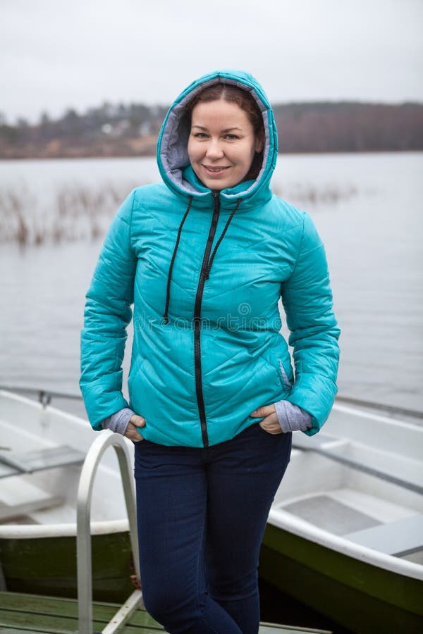 Mujer bastante caucásica en chaqueta azul con la capucha que se coloca en orilla del río debajo de la lluvia, mirando la cámara fotos de archivo Mujer bastante caucásica en chaqueta azul con la capucha que se coloca en orilla del río debajo de la lluvia, mirando la cámara fotos de archivo