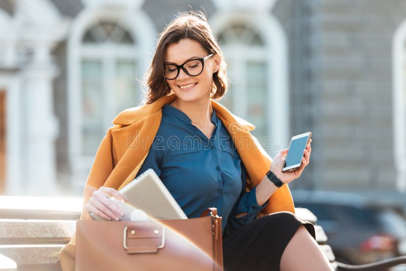 Mujer bonita sonriente en las lentes y la capa que sostienen el teléfono móvil fotografía de archivo libre de regalías Mujer bonita sonriente en las lentes y la capa que sostienen el teléfono móvil fotografía de archivo libre de regalías