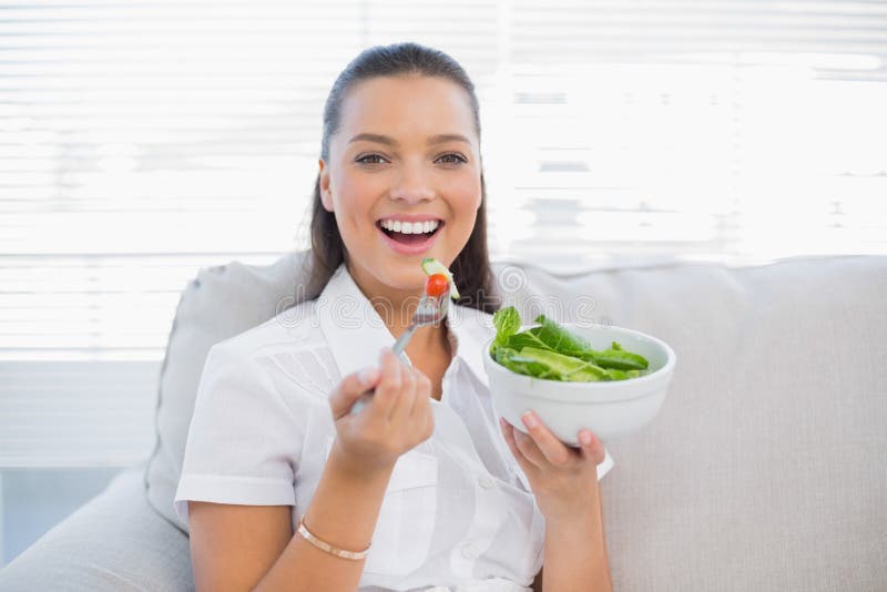 Mujer bonita sonriente que sostiene la ensalada sana que se sienta en el sofá imagen de archivo libre de regalías Mujer bonita sonriente que sostiene la ensalada sana que se sienta en el sofá imagen de archivo libre de regalías