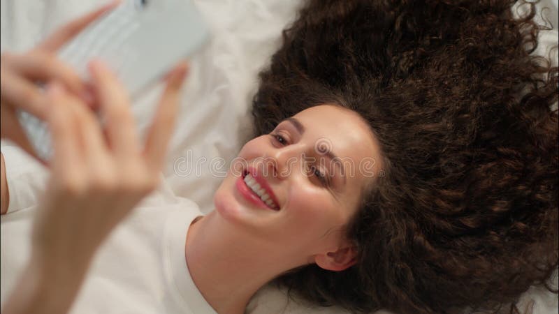 Mujer caucásica sonriente niña relajada sosteniendo el teléfono móvil en la cama despierta alegre mañana feliz usando un gadget de almacen de video