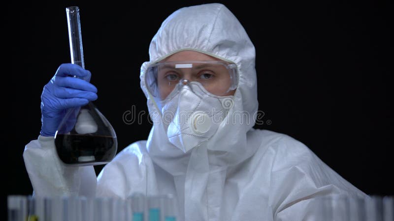 Mujer con traje protector estudiando frasco con lÃ­quido oscuro contra fondo negro