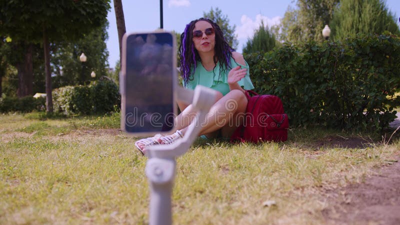 Mujer con un elegante peinado está sentada en el parque sobre el césped y grabando un video blog usando un smartphone. a almacen de metraje de vídeo