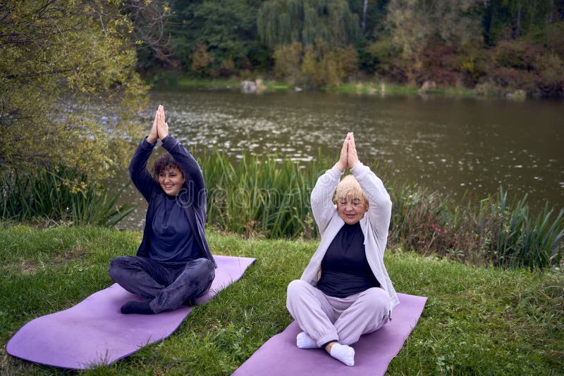Mujer de mediana edad enseña a su vieja madre ejercicios somáticos y yoga para mantener la salud cerca del río en el otoño foto de archivo