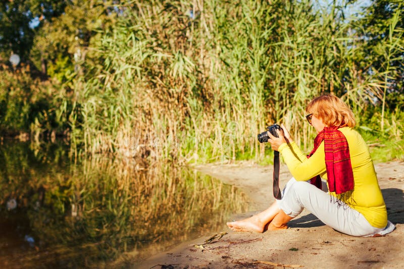 Mujer de mediana edad que comprueba imágenes en la cámara que se sienta por la orilla del río del otoño Mujer mayor que disfruta foto de archivo libre de regalías Mujer de mediana edad que comprueba imágenes en la cámara que se sienta por la orilla del río del otoño Mujer mayor que disfruta foto de archivo libre de regalías