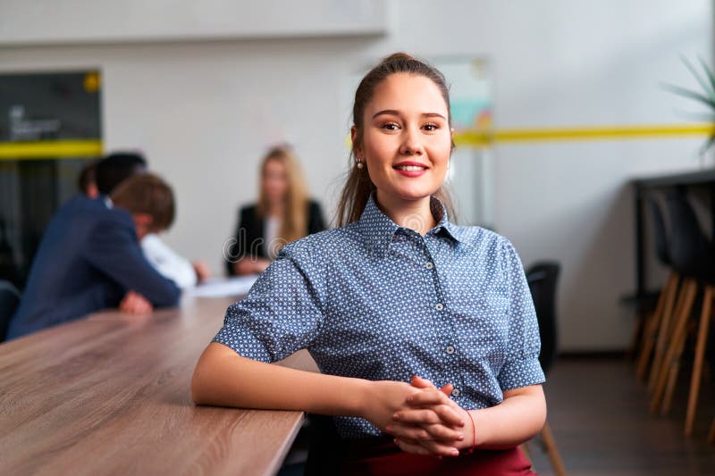 Modern workplace with team collaboration, leadership, and young female professional in focus. Confident Asian businesswoman sits at office desk, smiles at camera while colleagues work in background. Modern workplace with team collaboration, leadership, and young female professional in focus. Confident Asian businesswoman sits at office desk, smiles at camera while colleagues work in background.