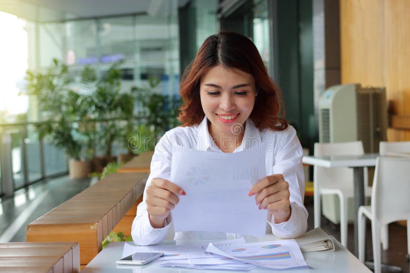 Mujer de negocios asiática feliz atractiva que sonríe con el papel del documento en su fondo de la oficina imagen de archivo Mujer de negocios asiática feliz atractiva que sonríe con el papel del documento en su fondo de la oficina imagen de archivo