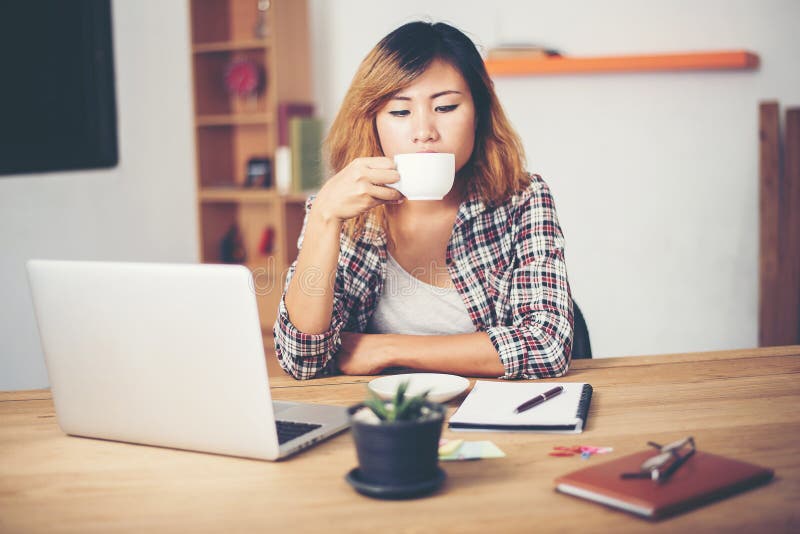 Mujer de negocios joven que se sienta en escritorio de oficina con la taza del café r fotos de archivo Mujer de negocios joven que se sienta en escritorio de oficina con la taza del café r fotos de archivo