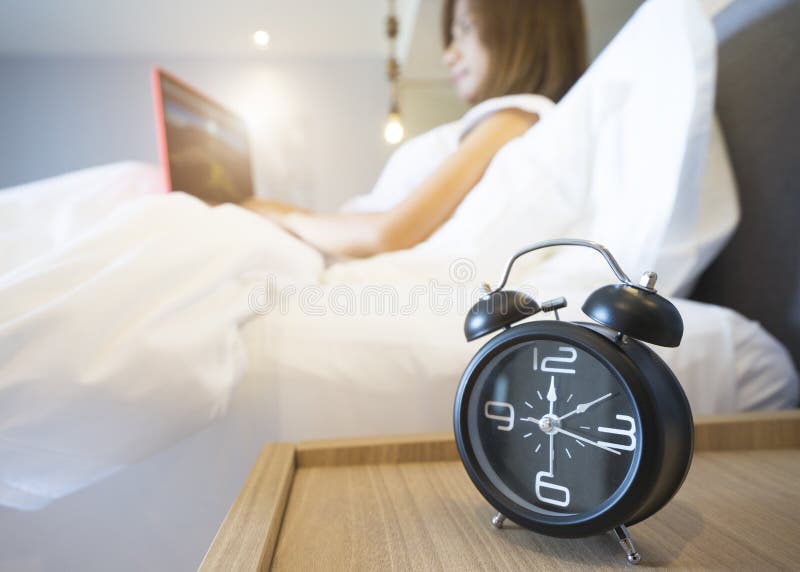 Mujer de negocios joven que se sienta en la cama blanca que trabaja en el ordenador portátil con el despertador en primero plano foto de archivo libre de regalías Mujer de negocios joven que se sienta en la cama blanca que trabaja en el ordenador portátil con el despertador en primero plano foto de archivo libre de regalías