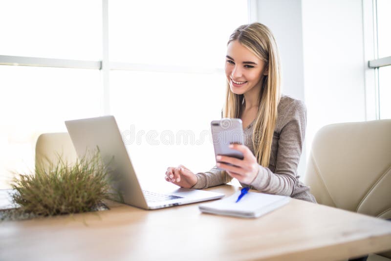 Mujer de negocios joven que usa el teléfono a disposición con el ordenador portátil en la oficina fotografía de archivo Mujer de negocios joven que usa el teléfono a disposición con el ordenador portátil en la oficina fotografía de archivo