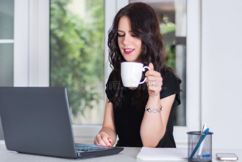Mujer de negocios joven y linda que trabaja en el ordenador portátil y que sostiene la taza de café imágenes de archivo libres de regalías Mujer de negocios joven y linda que trabaja en el ordenador portátil y que sostiene la taza de café imágenes de archivo libres de regalías
