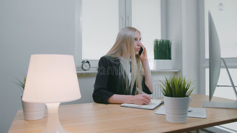 mujer de negocios que habla en smartphone en oficina Hembra rubia joven elegante en el traje de la oficina que se sienta en el lu