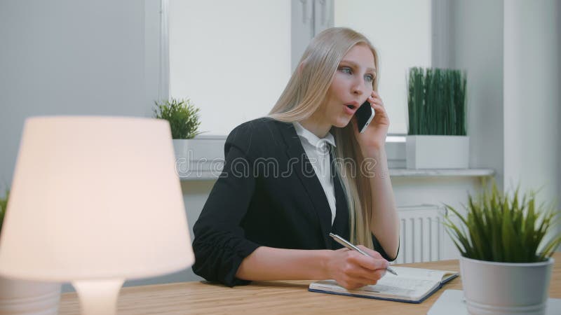 mujer de negocios que habla en smartphone en oficina Hembra rubia joven elegante en el traje de la oficina que se sienta en el lu