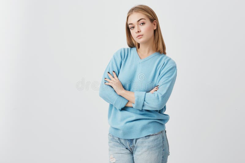 Portrait of fair-haired woman with warm dark eyes and healthy skin, standing with folded arms, looking directly into camera. Alluring girl with beautiful appearance dressed casually posing in studio. Beauty concept. Portrait of fair-haired woman with warm dark eyes and healthy skin, standing with folded arms, looking directly into camera. Alluring girl with beautiful appearance dressed casually posing in studio. Beauty concept