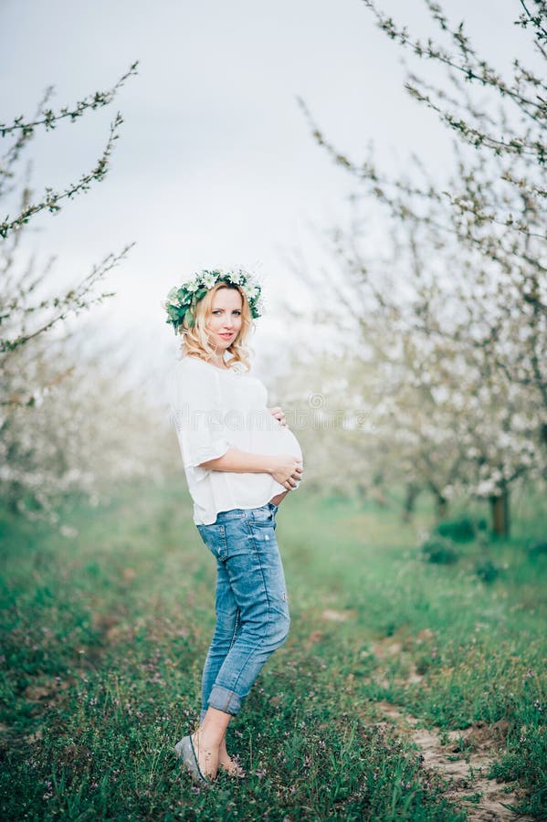 Mujer embarazada alegre joven hermosa en la guirnalda de flores en el vientre conmovedor principal mientras que camina en jardín fotos de archivo Mujer embarazada alegre joven hermosa en la guirnalda de flores en el vientre conmovedor principal mientras que camina en jardín fotos de archivo
