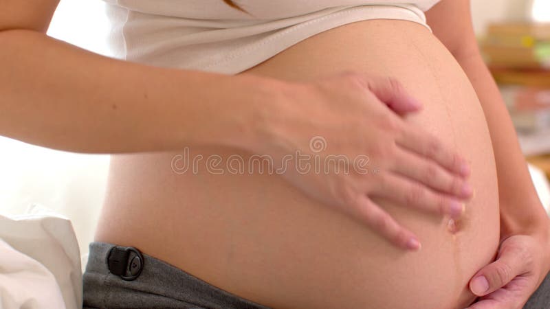 Mujer embarazada con la mano cerrada sentada en la cama sosteniendo y acariciando su gran barriga en casa embarazo de mujer joven almacen de metraje de vídeo