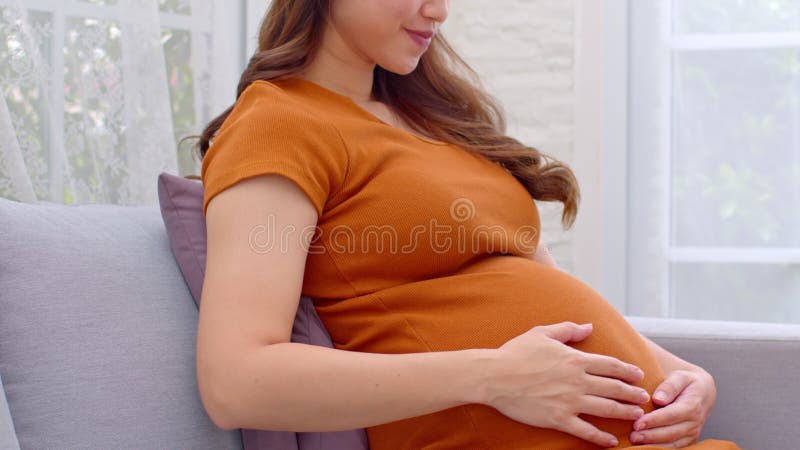 Mujer embarazada feliz lleva vestido marrón sentado en el sofá acariciando su gran barriga en casa embarazo joven disfrutando con almacen de video