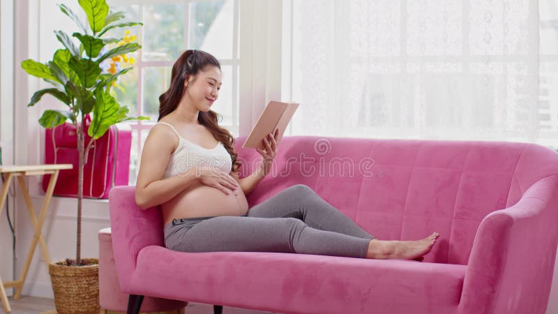 Mujer embarazada feliz sentada en el sofá sosteniendo un libro y leyendo para prepararse para cuidar el embarazo de bebé de una jo metrajes