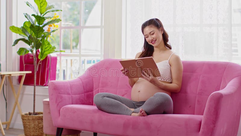 Mujer embarazada feliz sentada en el sofá sosteniendo un libro y leyendo para prepararse para cuidar el embarazo de bebé de una jo almacen de video