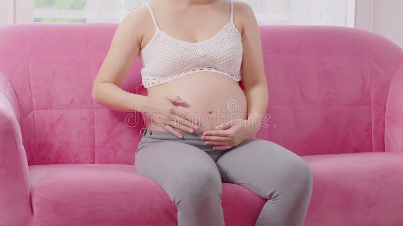Mujer embarazada feliz sentada en el sofá sosteniendo y acariciando su gran barriga en el acogedor embarazo en casa de una mujer j almacen de video