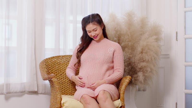 Mujer embarazada feliz sentada en el sofá sosteniendo y acariciando su gran barriga en el acogedor embarazo en casa de una mujer j almacen de video