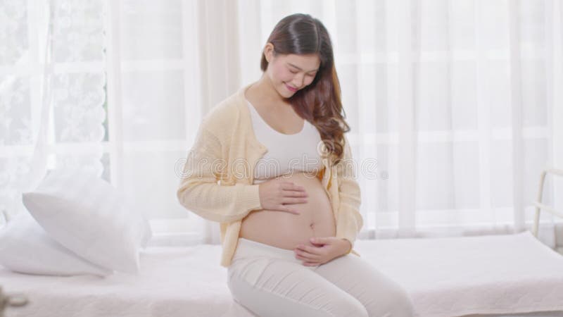 Mujer embarazada feliz sentada en la cama sosteniendo y acariciando su gran vientre en casa embarazo de una mujer joven disfrutand almacen de metraje de vídeo