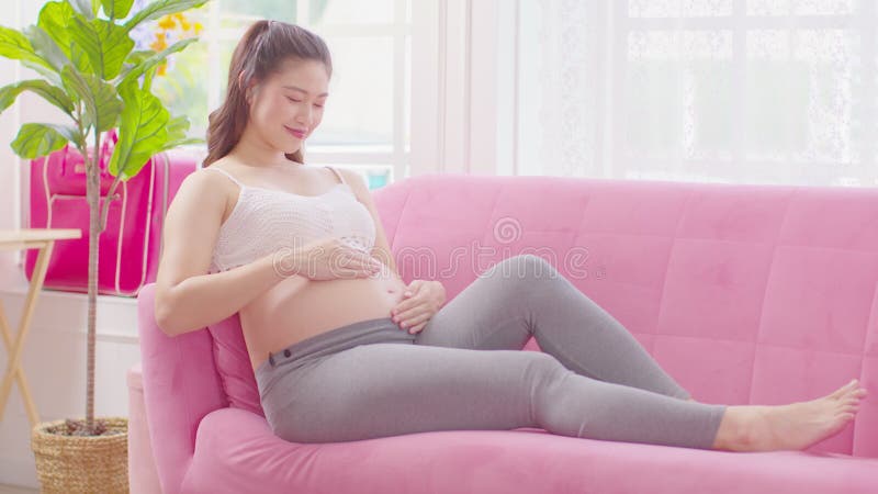 Mujer embarazada feliz sentada en un sofá rosa sosteniendo y acariciando su gran barriga en el acogedor embarazo en casa de una jo almacen de video
