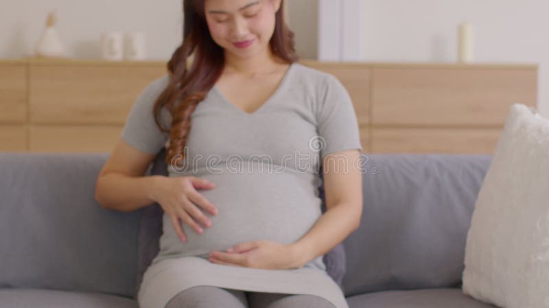 Mujer embarazada feliz sentada en un sofá sosteniendo y acariciando su gran vientre en el embarazo de su casa de una joven que dis metrajes