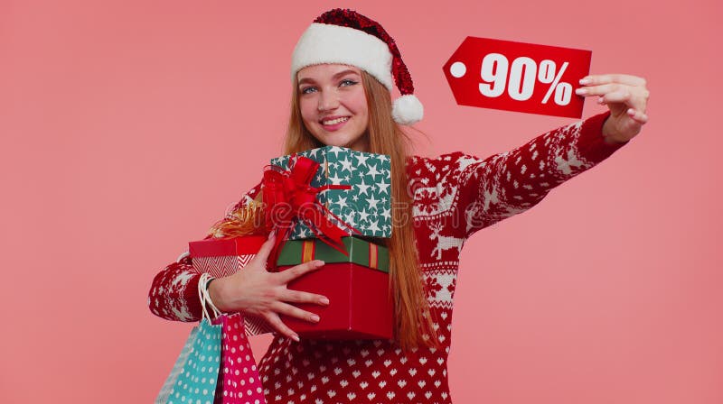 Adult girl wears red New Year sweater showing gift box and 90 Percent discount inscriptions banner text. Black Friday. Advertising low prices for shopping. Pink studio wall background. Merry Christmas. Adult girl wears red New Year sweater showing gift box and 90 Percent discount inscriptions banner text. Black Friday. Advertising low prices for shopping. Pink studio wall background. Merry Christmas