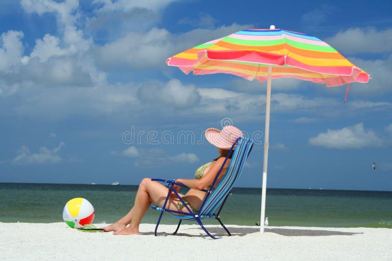 A woman sitting in a deck chair under colorful beach umbrella enjoying a day at the beach. Travel & Vacation Collection. A woman sitting in a deck chair under colorful beach umbrella enjoying a day at the beach. Travel & Vacation Collection.