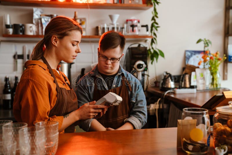 Mujer enseñando a un empleado con síndrome de Down a usar un terminal de pago en una cafetería fotografía de archivo