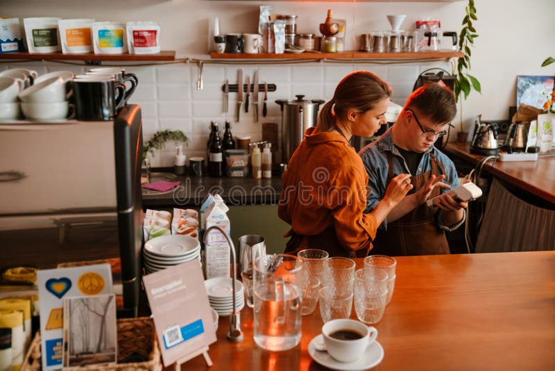 Mujer enseñando a un empleado con síndrome de Down a usar un terminal de pago en una cafetería imagen de archivo