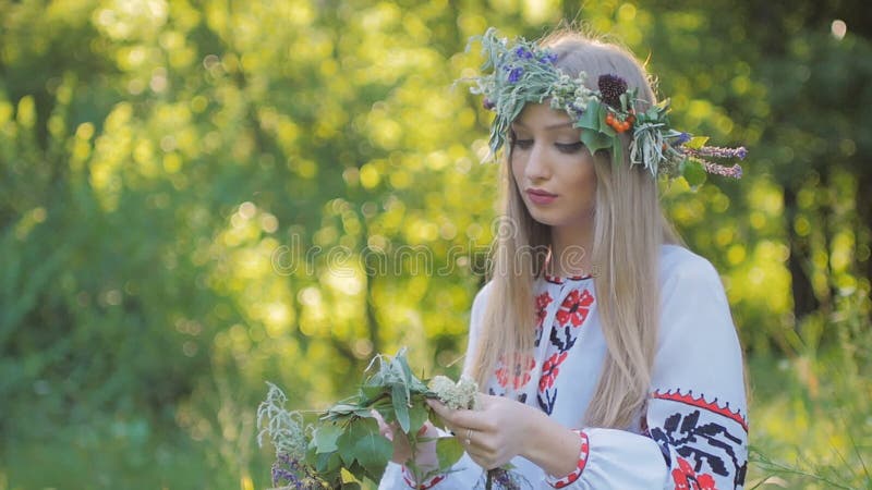 Mujer eslava con estampados de camisas bordadas teje una corona de flores silvestres sentada junto al fuego .