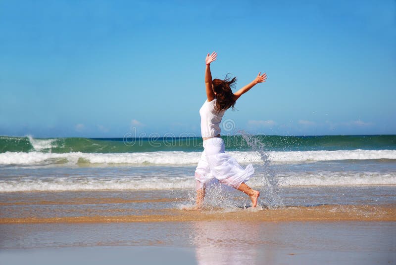 A Caucasian young woman dressed in white summer clothes running through the water on the beach raising up her arms to express her happiness. A Caucasian young woman dressed in white summer clothes running through the water on the beach raising up her arms to express her happiness.