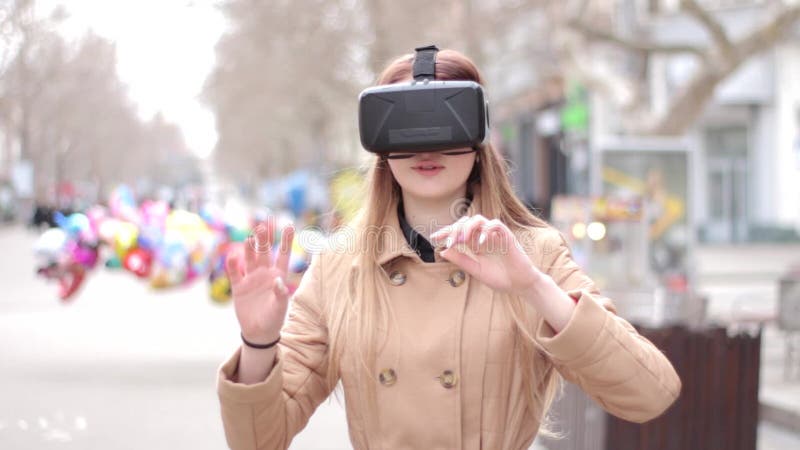 Mujer feliz joven en la calle en la capa beige que juega teniendo vidrios del ciberespacio de las auriculares de la realidad virt almacen de video Mujer feliz joven en la calle en la capa beige que juega teniendo vidrios del ciberespacio de las auriculares de la realidad virt almacen de video