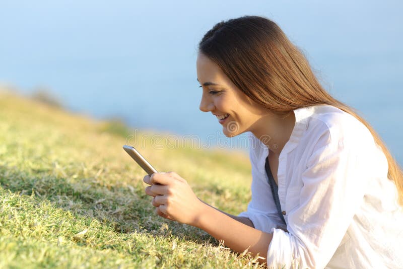 Mujer feliz que usa un smartphone que miente en la hierba imagen de archivo libre de regalías Mujer feliz que usa un smartphone que miente en la hierba imagen de archivo libre de regalías