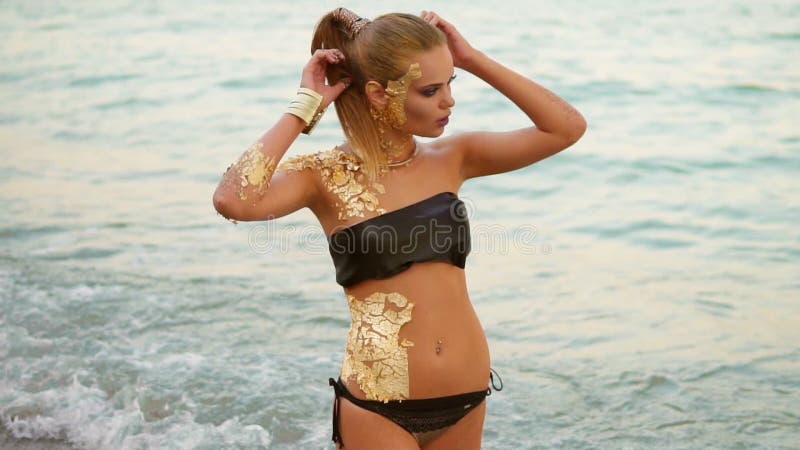 Mujer hermosa con el maquillaje de oro profesional que se coloca en el agua en la playa, mirando en la cámara atractivo