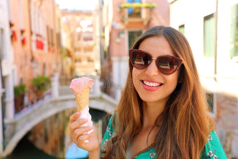 Mujer hermosa feliz con las gafas de sol que sostienen el helado del gelato en su mano y que sonríen en la cámara con el puente d foto de archivo Mujer hermosa feliz con las gafas de sol que sostienen el helado del gelato en su mano y que sonríen en la cámara con el puente d foto de archivo