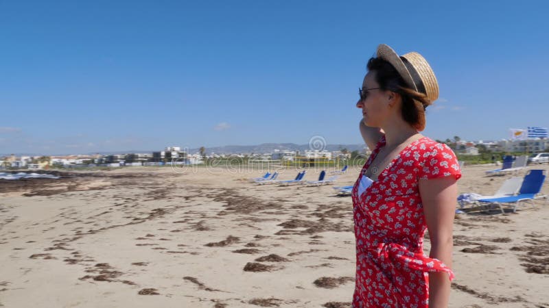 Mujer hermosa joven en la situación roja del vestido y del sombrero sola en la playa vacía con los sunbeds Playa de la arena con