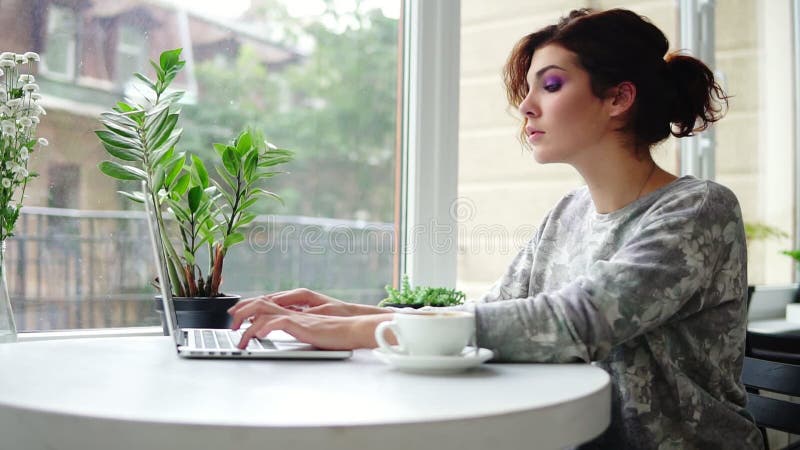 Mujer hermosa joven que se sienta cerca de la ventana en café y que trabaja en el ordenador portátil Muchacha que practica surf I