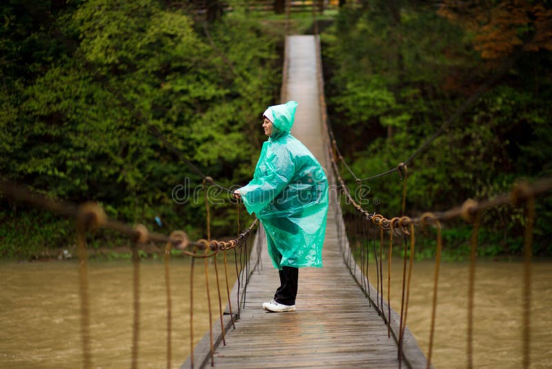 Mujer hermosa mayor del viajero en el río cruzado de la chaqueta azul de la lluvia por el puente con bisagras en el bosque, disfr fotos de archivo Mujer hermosa mayor del viajero en el río cruzado de la chaqueta azul de la lluvia por el puente con bisagras en el bosque, disfr fotos de archivo