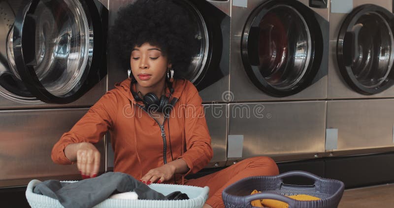 Mujer joven afroamericana feliz que clasifica el lavadero en la cesta que clasifica la ropa antes de la sentada que se lava en el