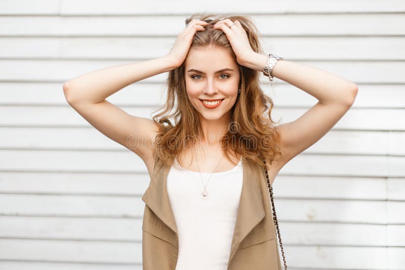 Mujer joven alegre en un chaleco beige del verano elegante en una camiseta elegante que presenta cerca de una pared blanca del vi fotografía de archivo libre de regalías Mujer joven alegre en un chaleco beige del verano elegante en una camiseta elegante que presenta cerca de una pared blanca del vi fotografía de archivo libre de regalías