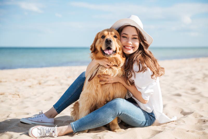 Mujer joven alegre que sienta y que abraza su perro en la playa fotografía de archivo Mujer joven alegre que sienta y que abraza su perro en la playa fotografía de archivo
