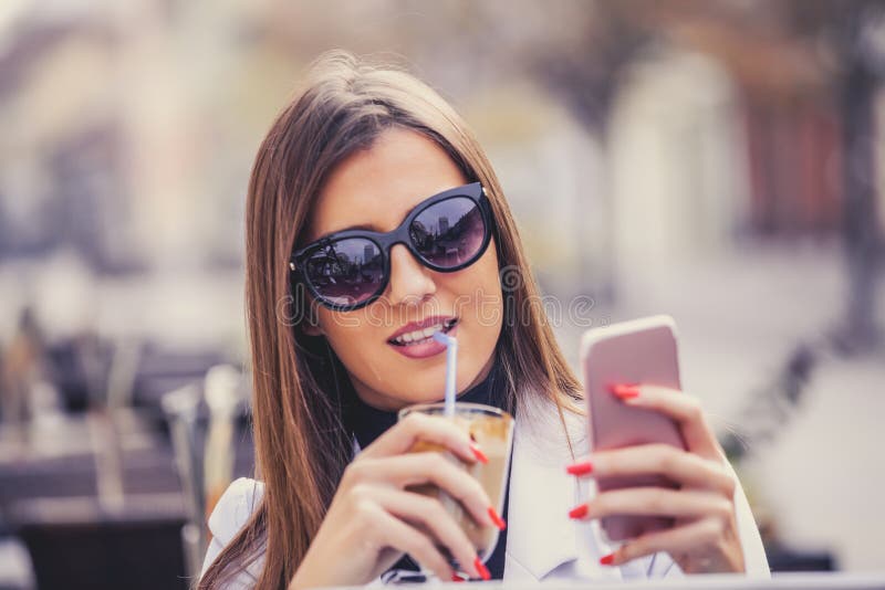 Mujer joven alegre que sostiene el teléfono elegante y que bebe choco caliente foto de archivo Mujer joven alegre que sostiene el teléfono elegante y que bebe choco caliente foto de archivo