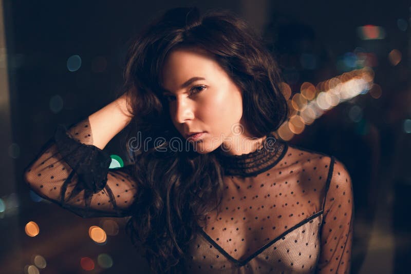 Mujer joven atractiva en vestido de cóctel negro delante de la ventana en la noche fotos de archivo Mujer joven atractiva en vestido de cóctel negro delante de la ventana en la noche fotos de archivo
