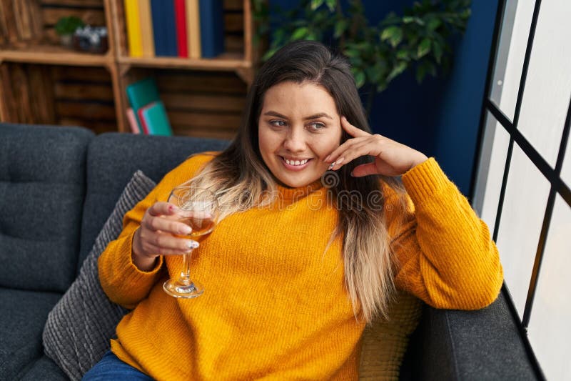 Mujer joven, bella y grande, tomando vino en el sofá de su casa foto de archivo libre de regalías
