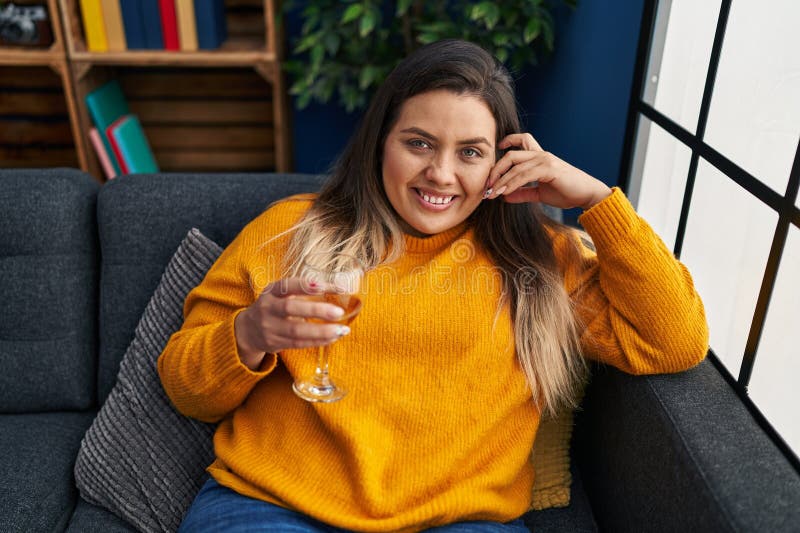 Mujer joven, bella y grande, tomando vino en el sofá de su casa fotos de archivo